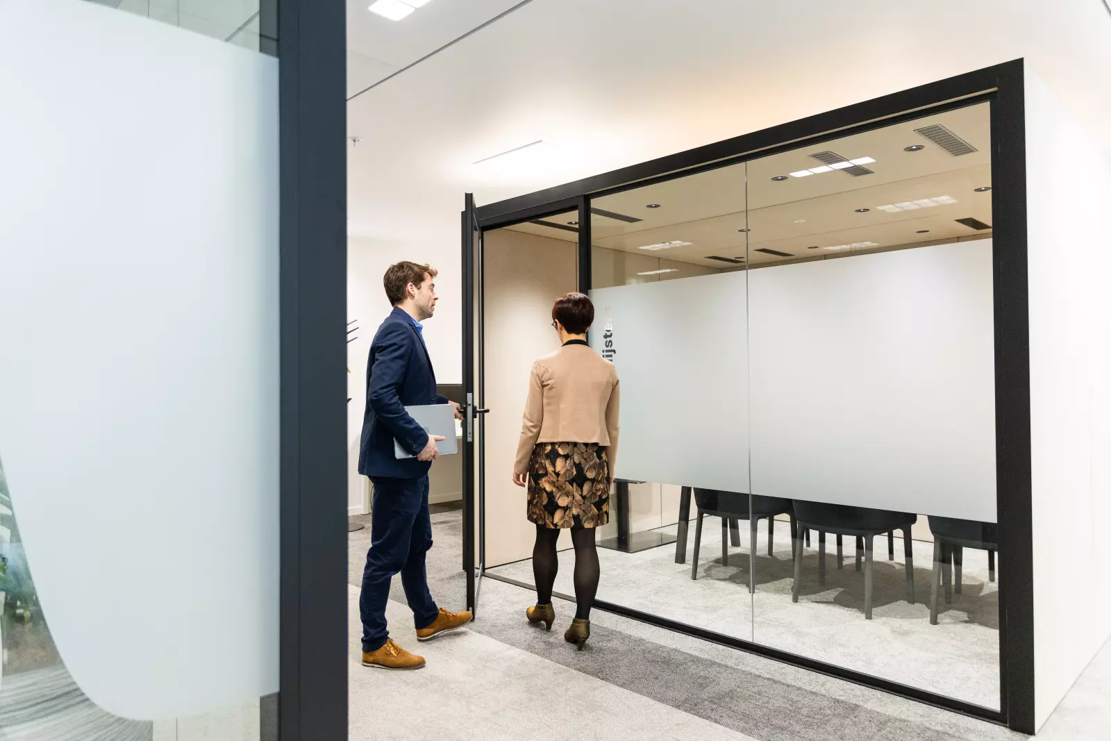 Deux collègues entrent dans une salle de réunion vitrée dans un bureau moderne.